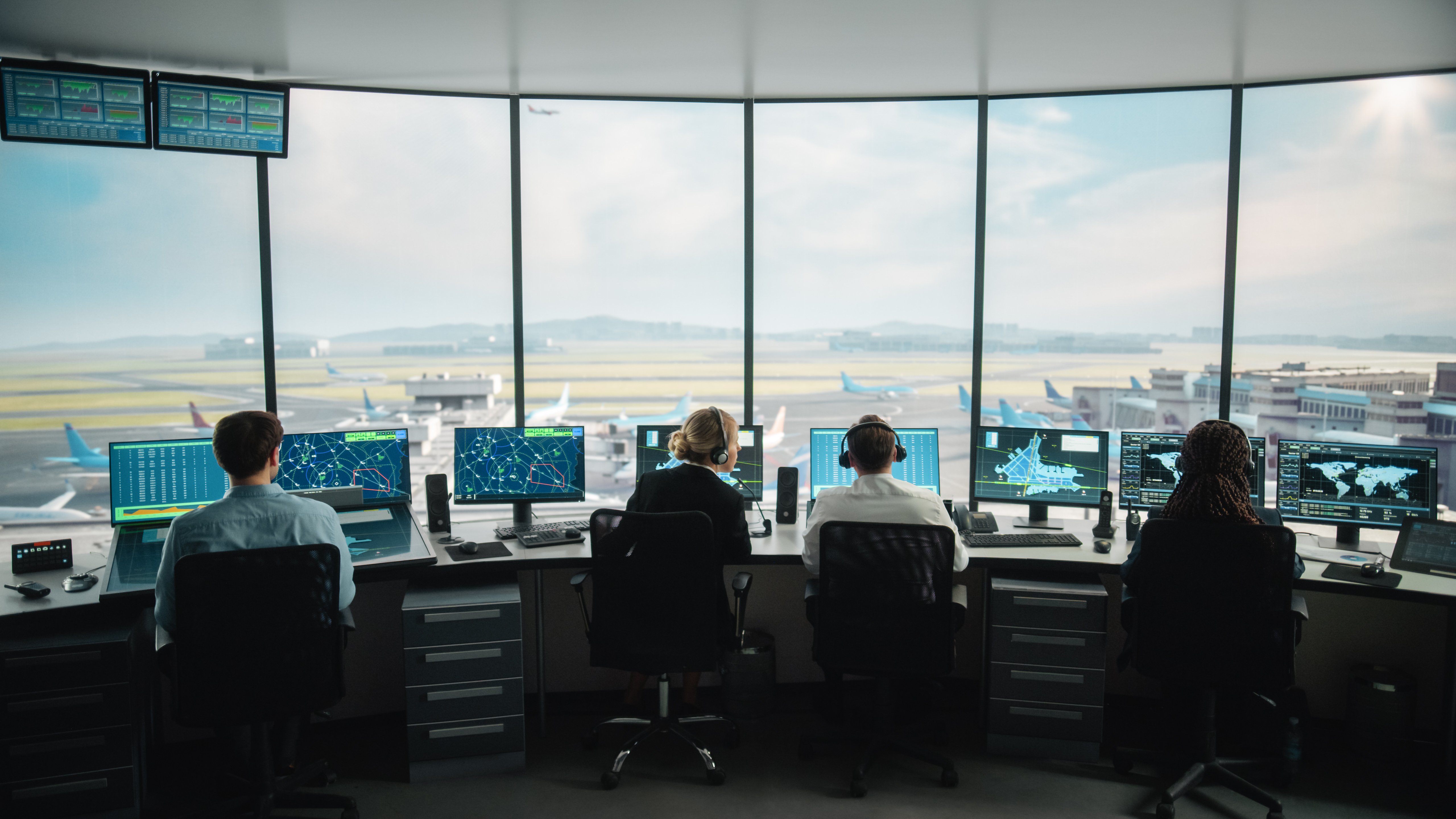 Cuatro controladores aéreos están sentados en sus puestos de trabajo en la torre de control de un aeropuerto, cada uno de ellos supervisando varias pantallas que muestran radares, trayectorias de vuelo, mapas del aeropuerto y datos globales. Las grandes ventanas panorámicas ofrecen una visión clara de la pista del aeropuerto, los aviones en rodaje y los edificios de la terminal. El equipo coordina los movimientos de los aviones en un entorno seguro y de alta tecnología.