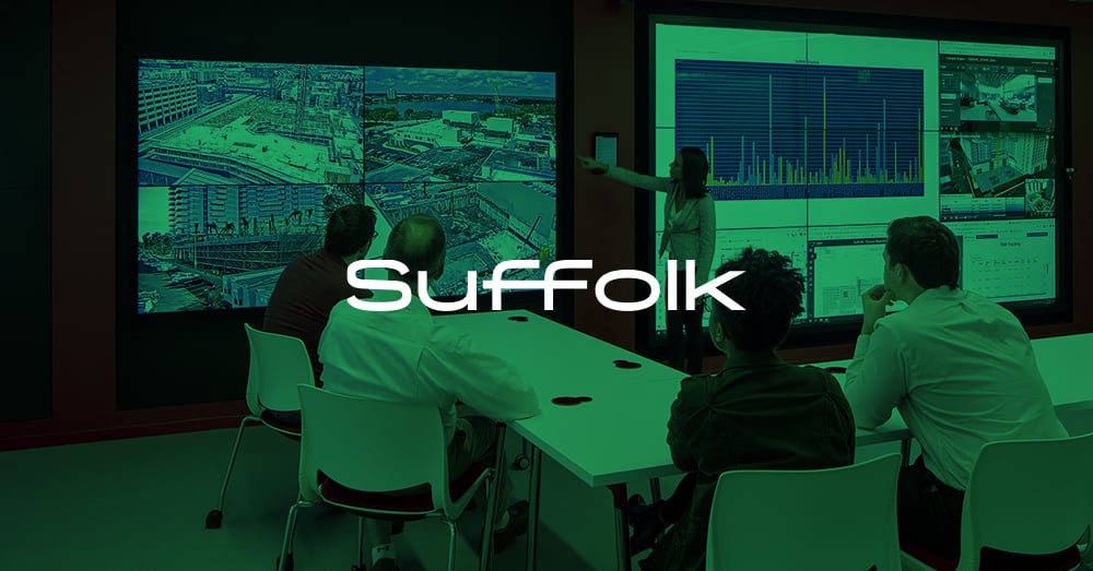 Un empleado de pie hace una presentación utilizando un videowall, 4 empleados ven la presentación sentados en una mesa en el L.A. Smart Lab de Suffolk con una superposición verde y el logotipo
