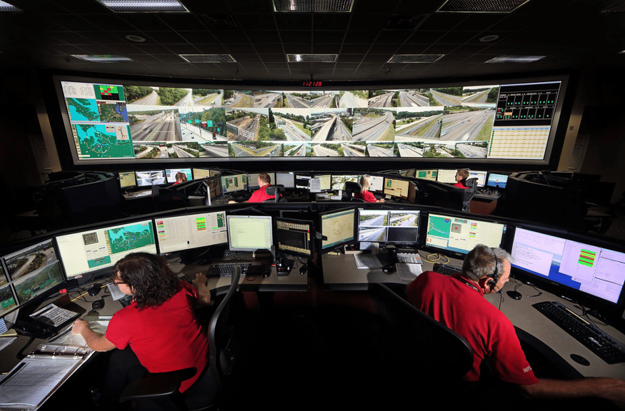 Un hombre y una mujer trabajan en sus puestos de trabajo en una sala de operaciones de seguridad, con una pared de vídeo detrás de ellos que muestra imágenes de cámaras de tráfico en directo