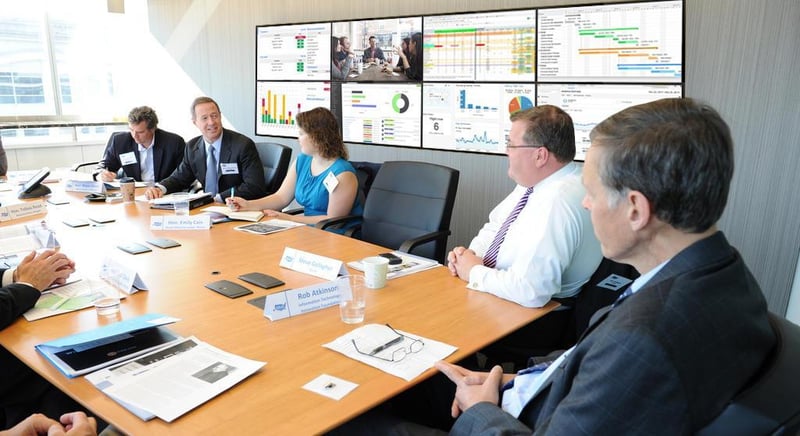 5 empleados discuten sentados en una mesa en una sala de reuniones con un videowall que muestra visualizaciones de datos
