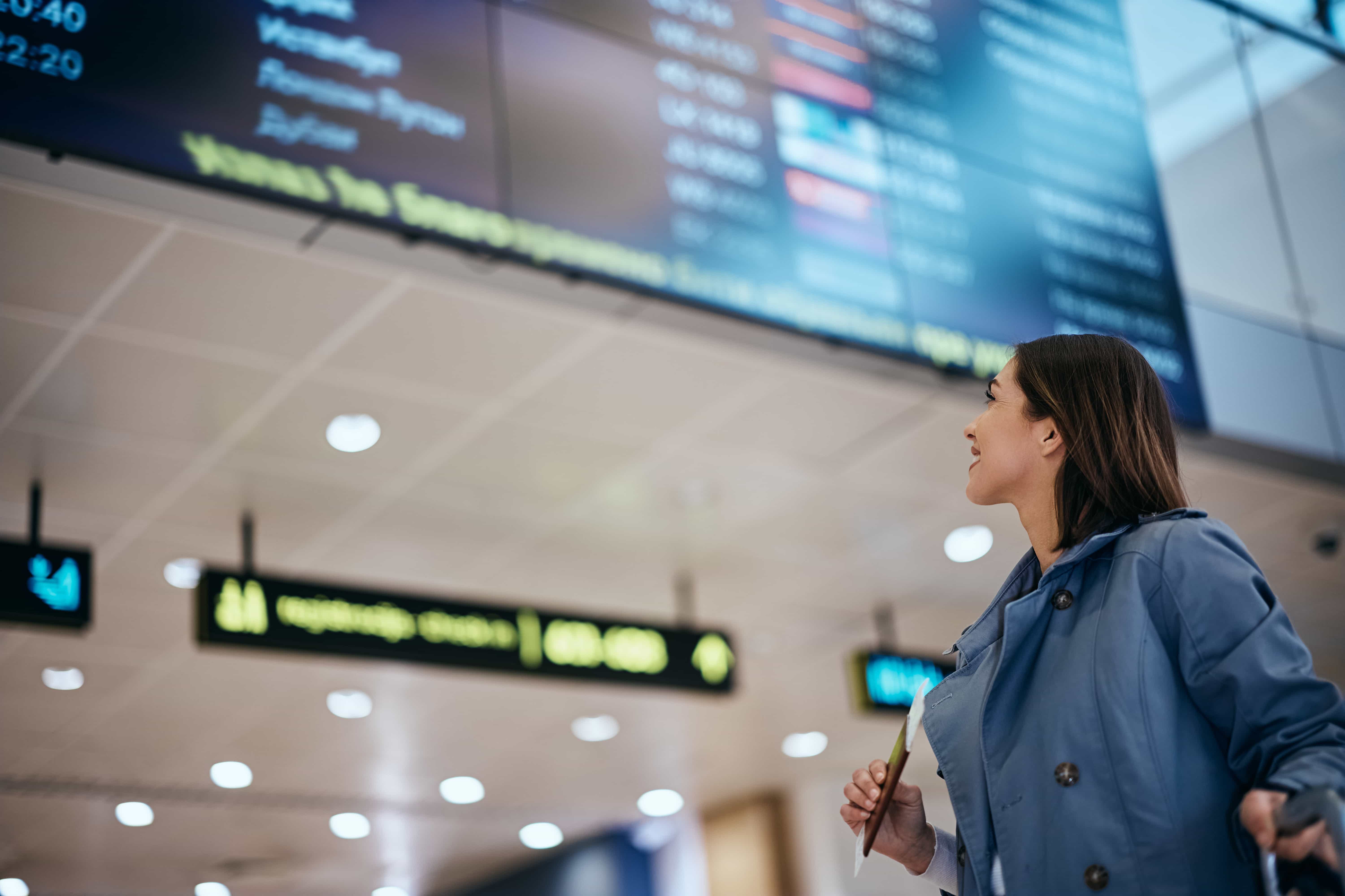  Una mujer con chaqueta azul claro y camisa blanca mira hacia un gran tablero de salidas en un aeropuerto. Sonríe ligeramente y sostiene lo que parece ser un pasaporte o un billete en la mano derecha. El panel de salidas, que está desenfocado, muestra diversa información sobre vuelos. El techo del aeropuerto con luces también es visible en el fondo.