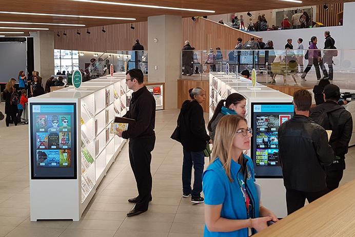 Many people in a library walking and viewing shelves with digital signage on them.