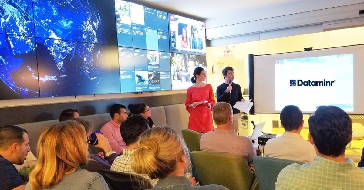 Empleados de Dataminr en una sala de reuniones sentados frente a un proyector y al lado de una pared de vídeo con medios sociales y noticias actualizadas