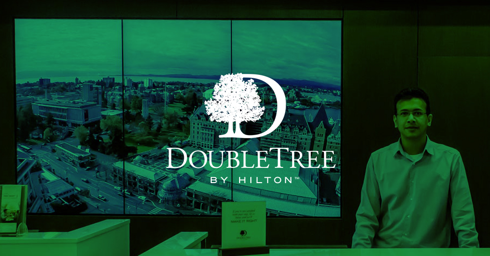 Recepcionista de DoubleTree by Hilton y pared de vídeo de 3 paneles detrás de él mostrando puntos de referencia de Victoria, Canadá, con superposición verde y logotipo