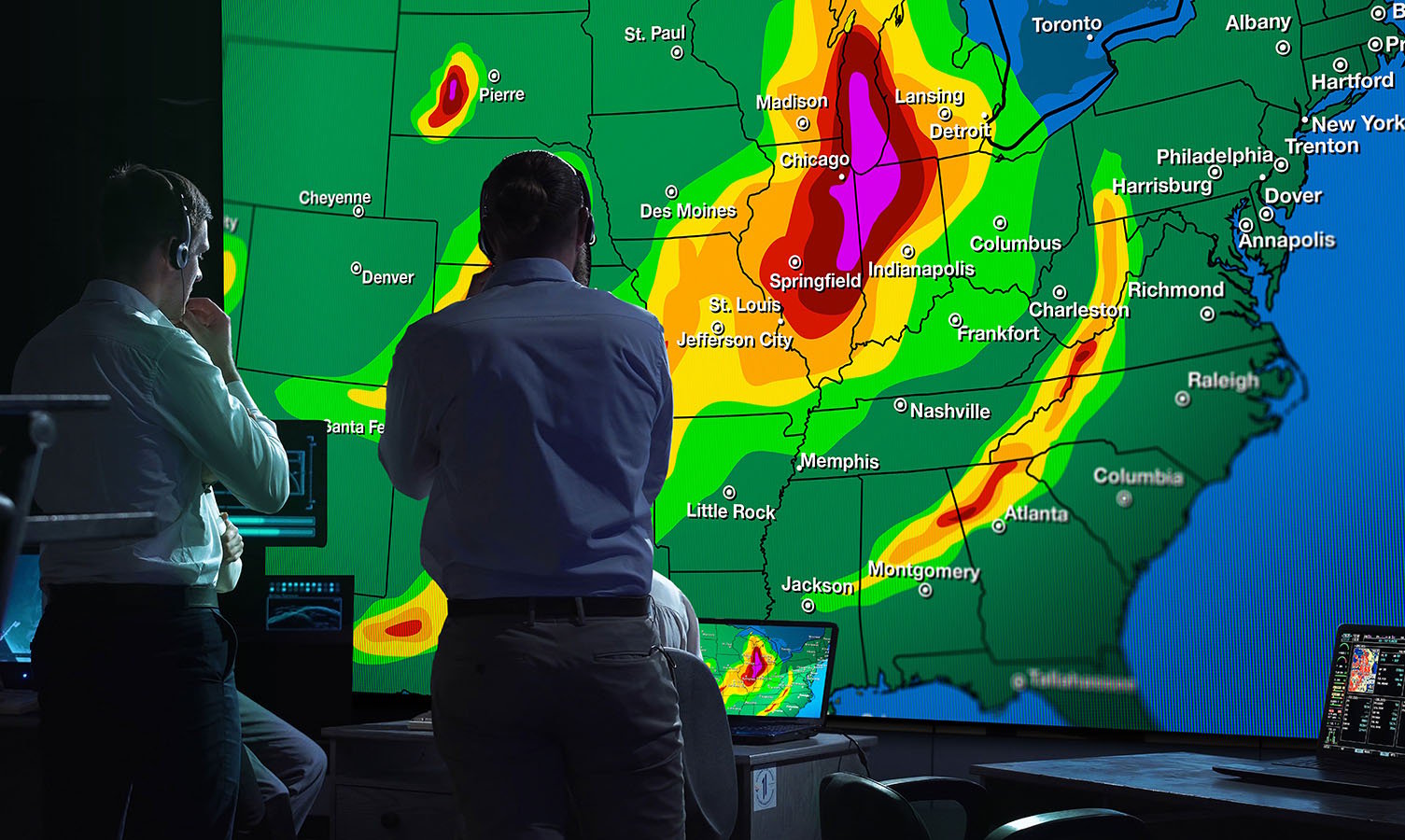 2 hombres en una sala de control meteorológico, mirando una gran pared de vídeo que muestra un mapa meteorológico de los EE.UU.