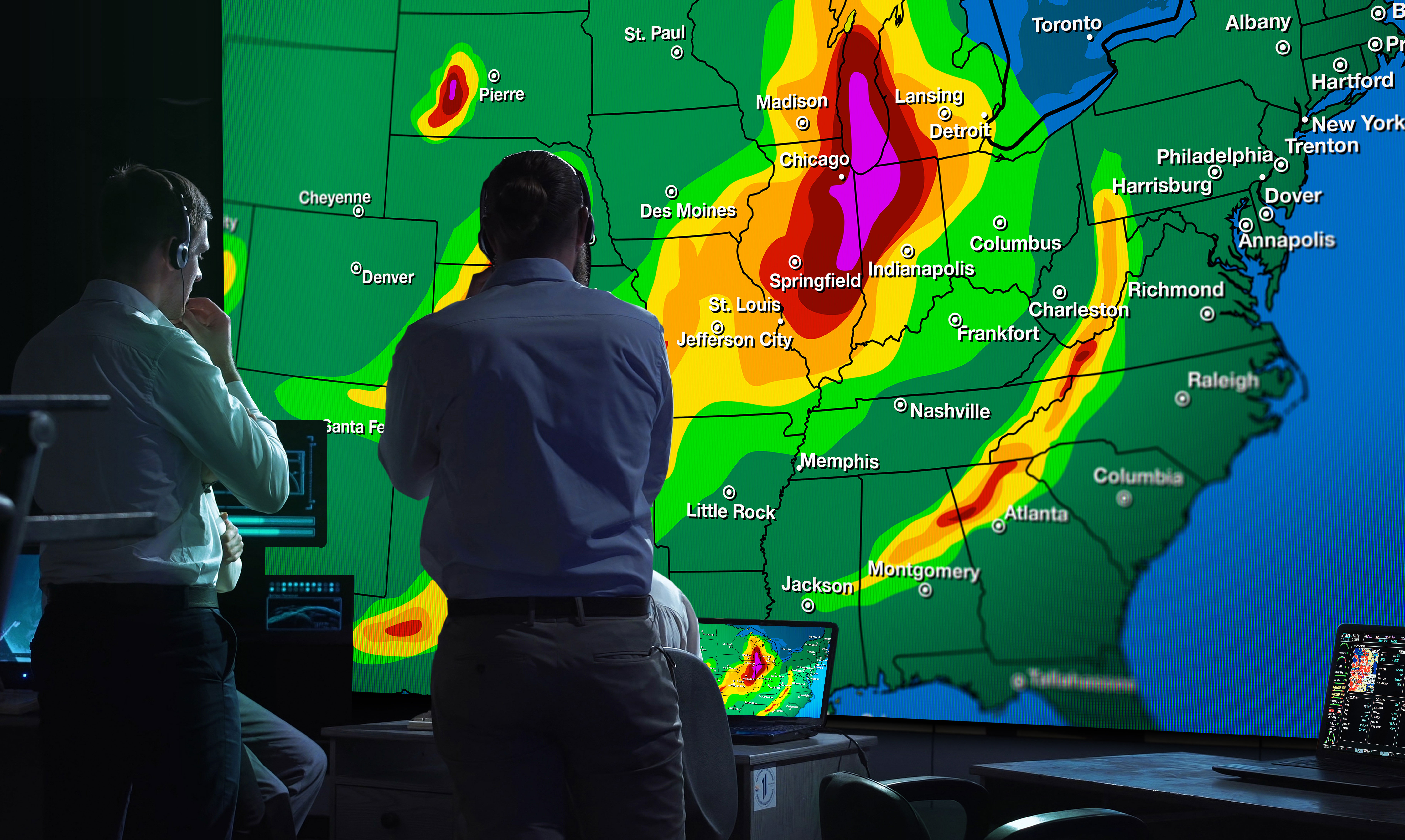 2 men in a weather monitoring control room, looking at a large video wall displaying a weather map of the USA