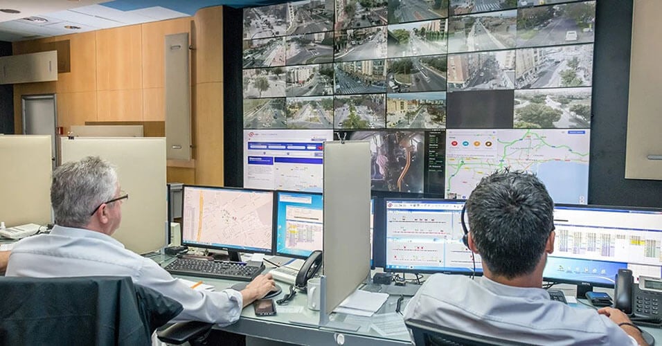 Dos empleados de la EMT que supervisan las operaciones de tránsito a través de sus puestos de trabajo y un muro de vídeo que muestra imágenes de cámaras en directo, mapas de tránsito y sitios web